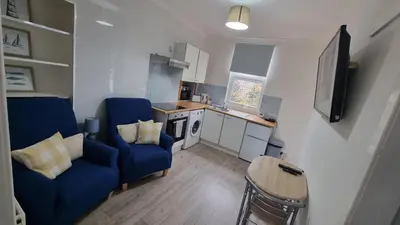 a living room with two blue chairs and a kitchen at Cove Bay 1 Bed Flat in Largs