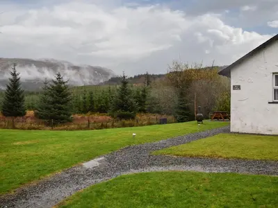 a white building in a field with a mountain in the background at Pine Cottage in Killin