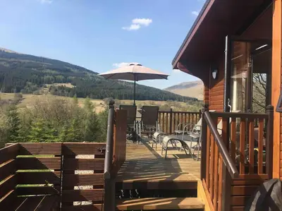 a wooden deck with a table and an umbrella at Teaghlach Luxury Lodge with hot tub Balquhidder Mhor Lodges in Lochearnhead