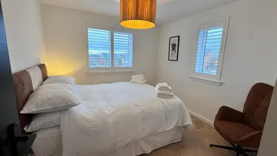 a bedroom with a bed and a chair and two windows at West Coast Home From Home in Stevenston