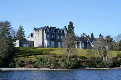 a large building on a hill next to a body of water at No 8 Lomond Castle in Luss