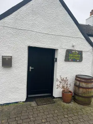 a white building with a black door and a barrel at Old Brewery Cottage - Ellenabeich, Easdale - Isle of Seil