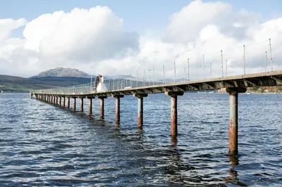 a bride and groom standing on a bridge over water at Rosslea Hall Hotel in Rhu