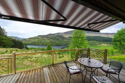 a deck with a table and chairs and a view of a lake at Kingfisher Lodge with sensational loch views in Crianlarich