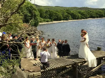 a bride and groom standing on a dock by the water at Loch Lomond shore Boat House in Balmaha