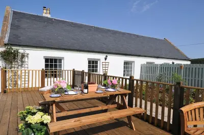 a wooden deck with a table and a white house at No 1 Ramageton at Carnell Estates in Hurlford
