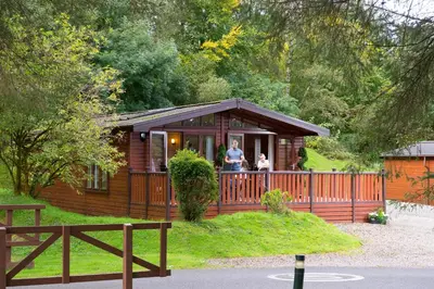 a wooden cabin with two people on the porch at Lomond Woods Holiday Park in Balloch