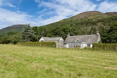 a house in a field with mountains in the background at Enchanting,rustic, Dundurn Mill with stunning views and private river in Comrie