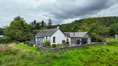 a white house with a stone wall in a field at Blair Cottage in Rowardennan