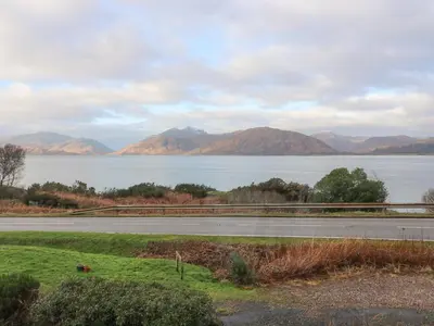 a road next to a body of water with mountains at Sea Otter Cottage in Appin