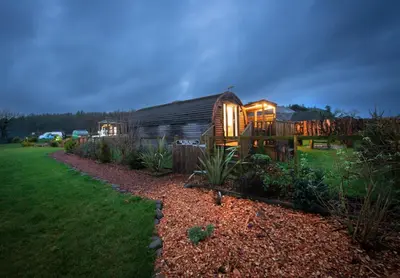 a large wooden cabin in a garden at night at The Galloway Steading - The Stag Hoose family cabin with Hot Tub and Fire Pit in Glenluce