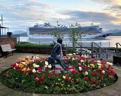 a statue of a woman in a flower garden with a cruise ship at clyde hub in Gourock