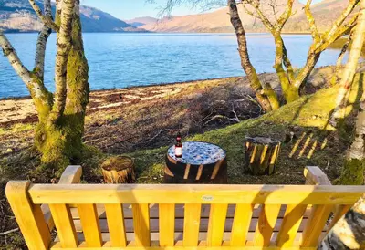a bench sitting next to a lake with a table at Seal Point Cabin - Luxury Glamping in Cairndow