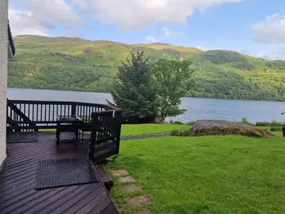 a deck with a picnic table and a view of a lake at WATERSIDE 3 BED COTTAGE, HOT TUB, SAUNA, PVT BEACH in Arrochar
