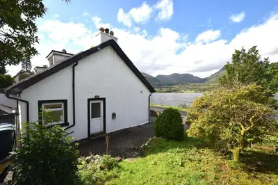 a white cottage with a lake in the background at 1 Planetree Cottage - Loch View in Lochgoilhead