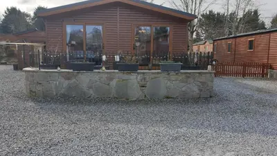 a log cabin with a stone wall in front of a house at Red Squirrel Lodge in Auchterarder